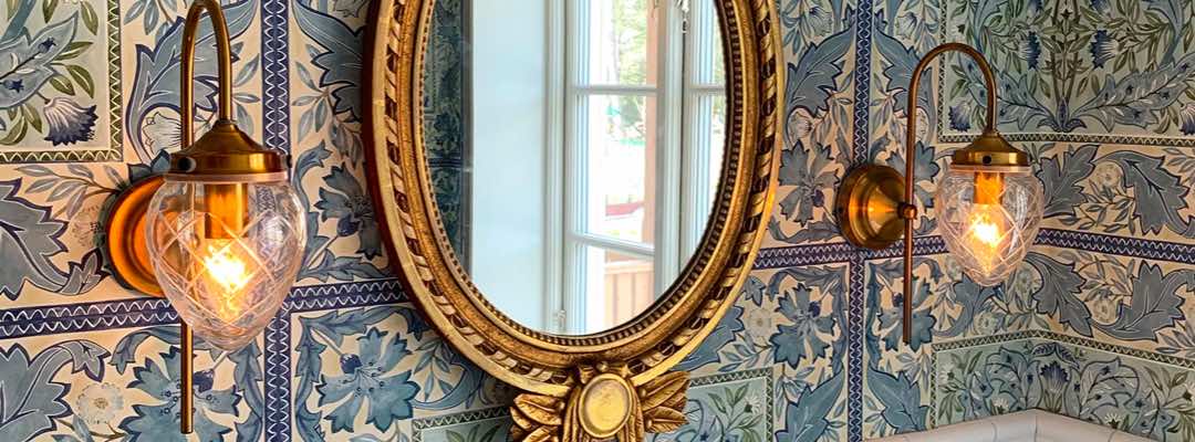 Bilden visar Badrumslampan Juni vägglampa i Slipat klarglas och Antik mässing, där två stycken sitter med en på vardera sida om spegeln i ett badrum.