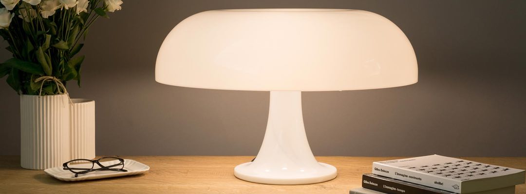 Bild på den vita Nesso bordslampan med rund, kupolformad lampskärm står på ett träbord bredvid en vas med vita blommor, ett par glasögon på ett fat och en stapel böcker.